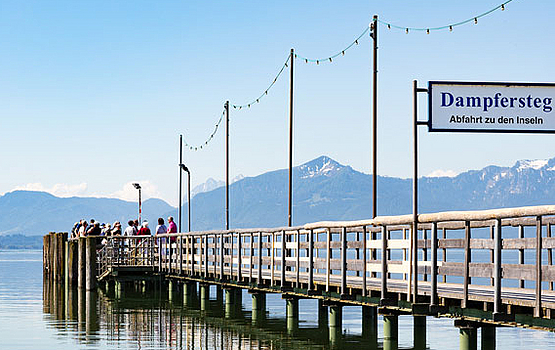 Langer Holzsteg mit Personen und Blick auf Wasser und Berge